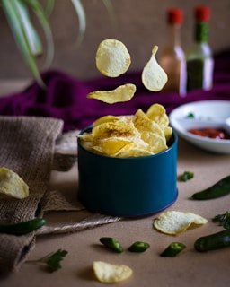 Crisp potato chips are scattered on and around a rustic surface, with several chips appearing to float above a teal container filled with more chips. Surrounding the scene are sliced green chili peppers and a small white dish containing a red sauce. Two bottles with red caps and blurred labels are placed in the background. A deep purple fabric adds contrast to the muted tones, and leafy plants are seen peeking in from the edges.