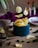 Crisp potato chips are scattered on and around a rustic surface, with several chips appearing to float above a teal container filled with more chips. Surrounding the scene are sliced green chili peppers and a small white dish containing a red sauce. Two bottles with red caps and blurred labels are placed in the background. A deep purple fabric adds contrast to the muted tones, and leafy plants are seen peeking in from the edges.