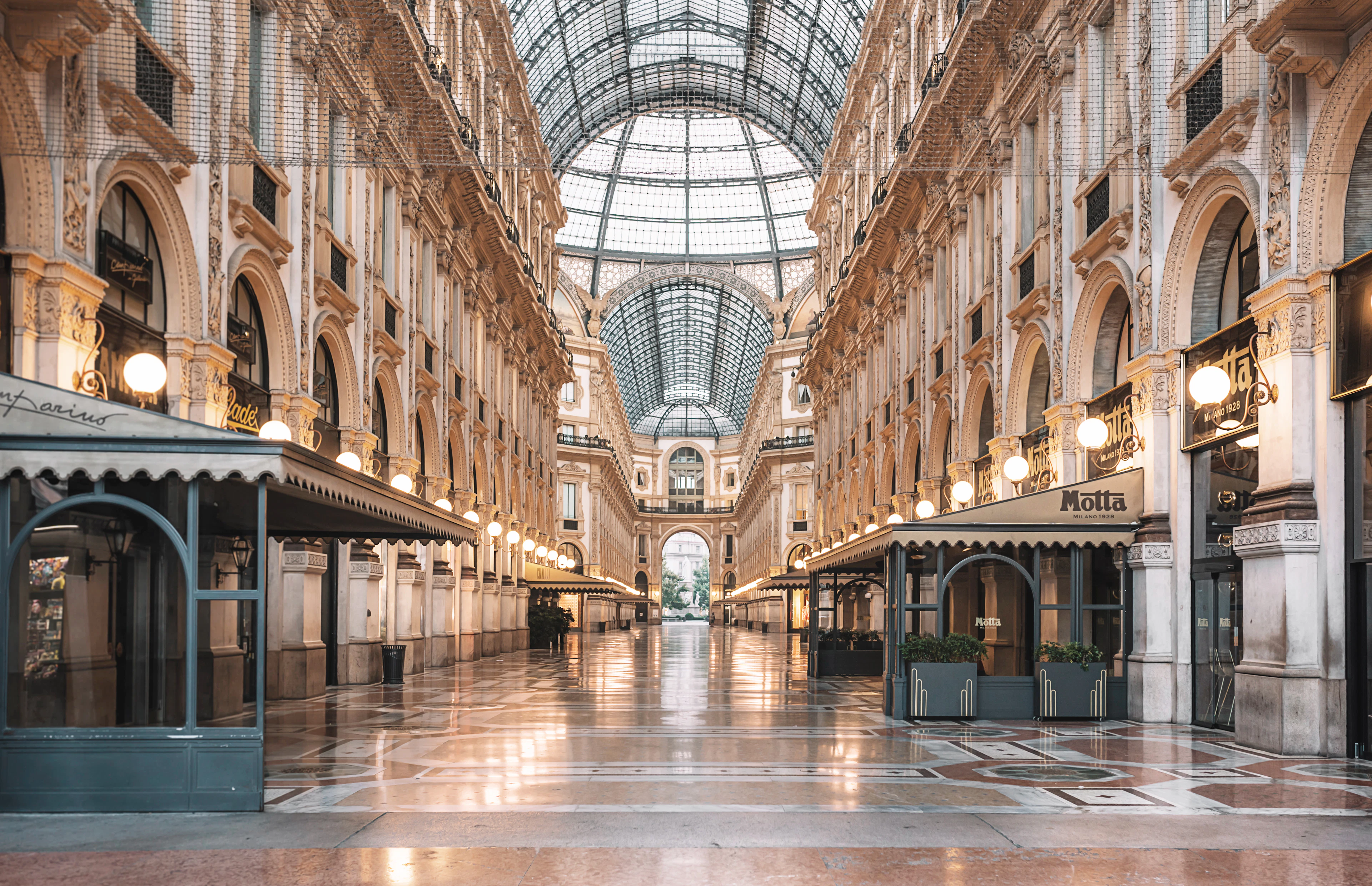 The marble arcade of Milan's Galleria Vittorio Emanuele II under its glass barrel vault, lamps glowing over the cafe awnings and a polished mosaic floor.