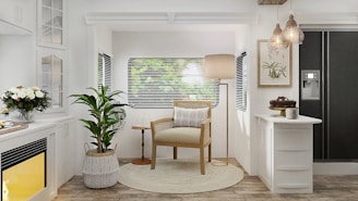 A cozy, well-lit living room area with a natural and minimalist aesthetic. There is a wooden armchair with a cushion placed on a circular rug in the center. A tall floor lamp is positioned behind the chair. Adjacent to the chair, a potted plant stands to the left, and a small wooden side table is on the right. The background features a window with blinds, through which blurred greenery is visible outside. On the left, there is a white fireplace with a yellow glow and a bouquet of flowers on the mantel. On the right, there is a kitchen counter with a cake and a coffee mug, and a black refrigerator with a water dispenser. Hanging pendant lights add a warm, welcoming glow to the room.