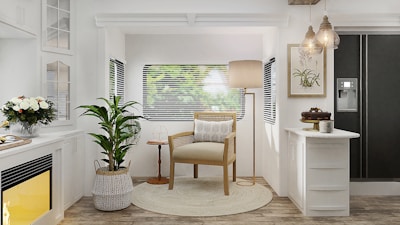 A cozy, well-lit living room area with a natural and minimalist aesthetic. There is a wooden armchair with a cushion placed on a circular rug in the center. A tall floor lamp is positioned behind the chair. Adjacent to the chair, a potted plant stands to the left, and a small wooden side table is on the right. The background features a window with blinds, through which blurred greenery is visible outside. On the left, there is a white fireplace with a yellow glow and a bouquet of flowers on the mantel. On the right, there is a kitchen counter with a cake and a coffee mug, and a black refrigerator with a water dispenser. Hanging pendant lights add a warm, welcoming glow to the room.
