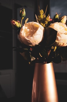 An elegant arrangement of blooming white roses with closed buds, placed in a tall, metallic vase. The lighting creates dramatic shadows on the petals and leaves, adding depth and texture to the flowers.