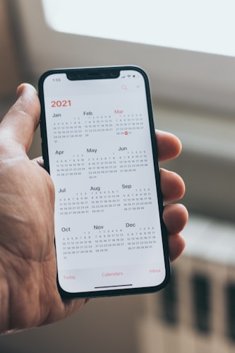 A hand holding a smartphone displaying a full-year calendar for 2021. The months and corresponding dates are clearly visible with a minimalist design. The background is blurred, highlighting the focus on the phone screen.