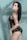 A person with long dark hair poses against a light blue background with shadow patterns of leaves. They are wearing a black leather outfit with fishnet stockings and chain accessories.