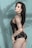 A person with long dark hair poses against a light blue background with shadow patterns of leaves. They are wearing a black leather outfit with fishnet stockings and chain accessories.