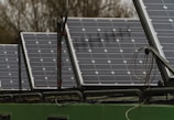 Several solar panels are arranged in a row on a structure, with visible wiring and mounts. The panels have a grid-like pattern with multiple solar cells. In the background, there are blurred trees indicating an outdoor setting.