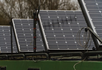 Several solar panels are arranged in a row on a structure, with visible wiring and mounts. The panels have a grid-like pattern with multiple solar cells. In the background, there are blurred trees indicating an outdoor setting.