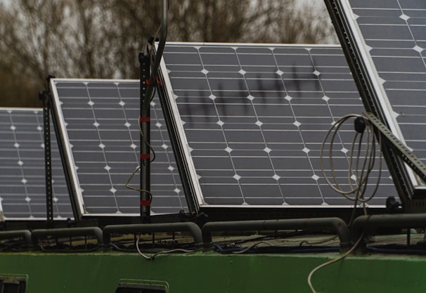 Several solar panels are arranged in a row on a structure, with visible wiring and mounts. The panels have a grid-like pattern with multiple solar cells. In the background, there are blurred trees indicating an outdoor setting.