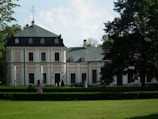 A large, elegant mansion with a green roof and multiple chimneys is surrounded by lush greenery and manicured lawns. The building features ornate architectural details and tall windows. Two fountains are placed symmetrically in front of the mansion, and a few people can be seen enjoying the weather in the garden.