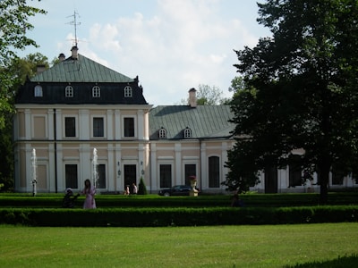 A large, elegant mansion with a green roof and multiple chimneys is surrounded by lush greenery and manicured lawns. The building features ornate architectural details and tall windows. Two fountains are placed symmetrically in front of the mansion, and a few people can be seen enjoying the weather in the garden.