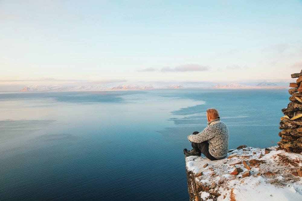 woman in brown knit cap and brown jacket sitting on rock near body of water during