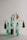 Three green plastic bottles are arranged on a white surface, each containing wilted flowers. The bottles have white labels with black text, highlighting environmental messages about plastic pollution. The setup is minimalistic, with a muted background.