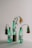 Three green plastic bottles are arranged on a white surface, each containing wilted flowers. The bottles have white labels with black text, highlighting environmental messages about plastic pollution. The setup is minimalistic, with a muted background.