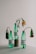 Three green plastic bottles are arranged on a white surface, each containing wilted flowers. The bottles have white labels with black text, highlighting environmental messages about plastic pollution. The setup is minimalistic, with a muted background.