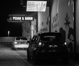 Two cars are parked in a dimly lit drive-thru area at night. A sign above displays 'SOSIS BAKAR, Try Me!' and another sign reads 'PESAN & BAYAR,' indicating an order and pay point. Murals of palm trees and animals decorate the walls.