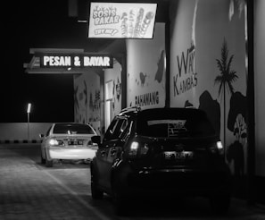 Two cars are parked in a dimly lit drive-thru area at night. A sign above displays 'SOSIS BAKAR, Try Me!' and another sign reads 'PESAN & BAYAR,' indicating an order and pay point. Murals of palm trees and animals decorate the walls.