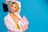 A person with short, curly blond hair poses against a bright blue background. They wear a light pink blazer over a yellow top and have vibrant makeup with pink blush and blue eyeshadow. In one hand, they hold a black cosmetic palette, while the other hand displays an open pink makeup case revealing various beauty products.