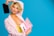 A person with short, curly blond hair poses against a bright blue background. They wear a light pink blazer over a yellow top and have vibrant makeup with pink blush and blue eyeshadow. In one hand, they hold a black cosmetic palette, while the other hand displays an open pink makeup case revealing various beauty products.