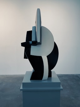 A modern abstract sculpture composed of geometric shapes is displayed on a pedestal. The piece combines smooth and angular lines, creating a striking contrast between black and white surfaces. The setting is minimalistic, with a plain wall and concrete floor providing a neutral backdrop that enhances the sculpture's presence.