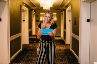 A woman stands in a well-lit hallway, holding a blue item that resembles a mirror or makeup palette. She appears to be applying makeup, possibly lipstick, with a focused expression. The hallway is elegant, with soft lighting and a chandelier.