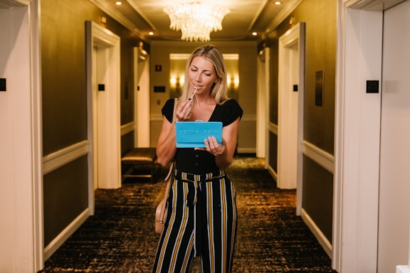 A woman stands in a well-lit hallway, holding a blue item that resembles a mirror or makeup palette. She appears to be applying makeup, possibly lipstick, with a focused expression. The hallway is elegant, with soft lighting and a chandelier.