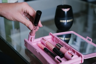 A hand holds a tube of lipstick or lip gloss over a pink makeup case containing several makeup items. The case is open on a reflective glass surface. In the background, a black cup with lip prints and the words 'MISS SWISS' is visible.