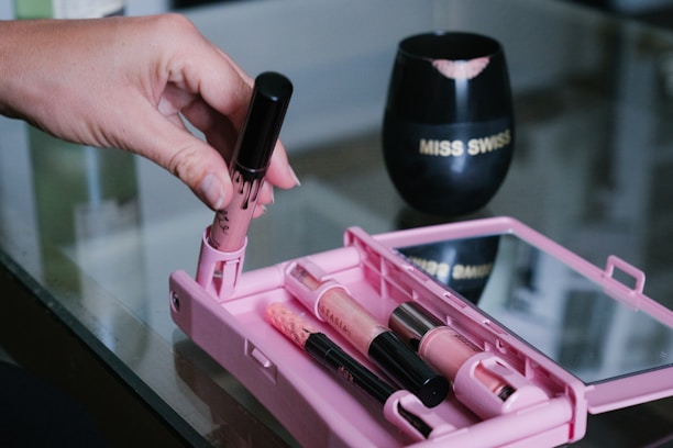 A hand holds a tube of lipstick or lip gloss over a pink makeup case containing several makeup items. The case is open on a reflective glass surface. In the background, a black cup with lip prints and the words 'MISS SWISS' is visible.