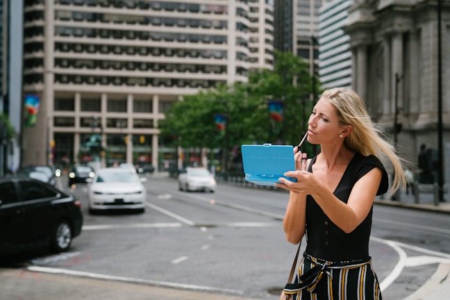 A stylish woman applying lipstick in front of a large urban window, city skyline blurred behind.