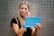 A person with long blonde hair is applying makeup using a blue folding mirror labeled 'MISS SWISS'. They hold the mirror in one hand and a makeup tool in the other. The background features a gray, perforated wall.