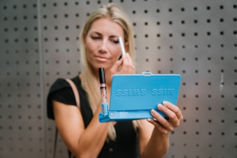 A person with long blonde hair is applying makeup using a blue folding mirror labeled 'MISS SWISS'. They hold the mirror in one hand and a makeup tool in the other. The background features a gray, perforated wall.