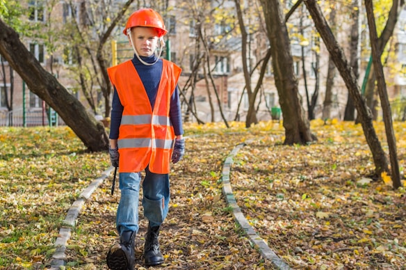 A young child wearing a bright orange safety vest and helmet walks on a narrow path surrounded by trees with fallen leaves. The child is also wearing gloves, a blue long-sleeve shirt, jeans, and black boots. In the background, there are residential buildings partly visible through the trees.