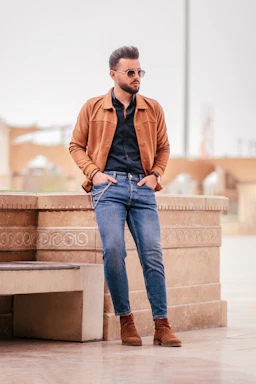 man in brown jacket and blue denim jeans sitting on brown concrete bench during daytime
