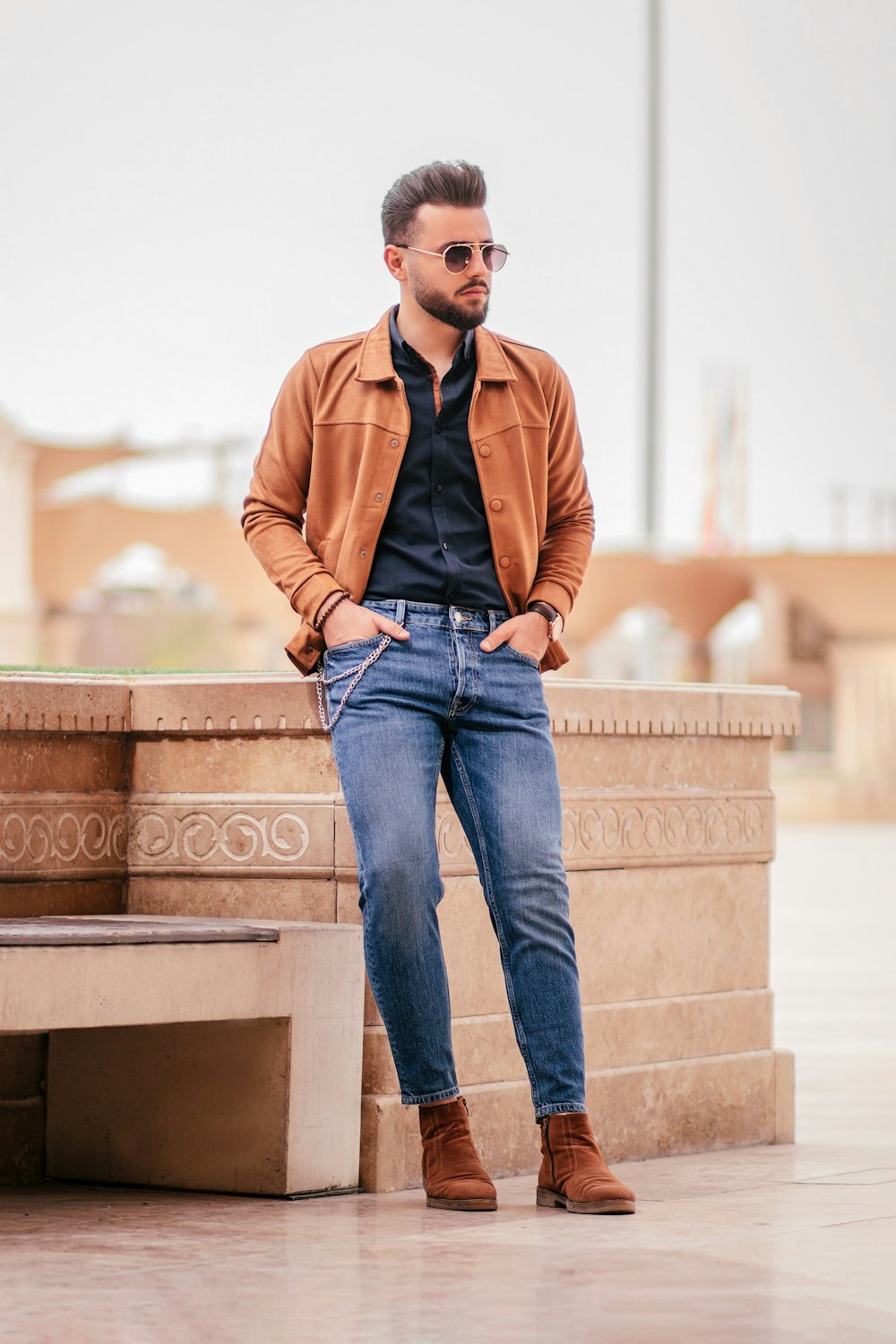 uomo in giacca marrone e jeans blu denim seduto su panchina di cemento marrone durante il giorno