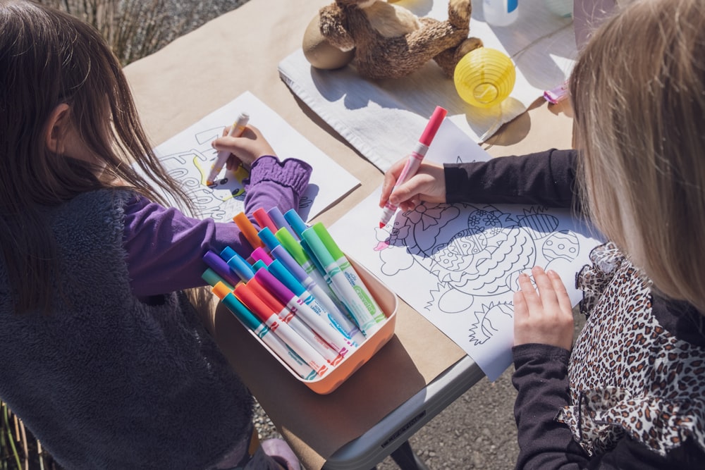 Dos niñas sentadas en una mesa con marcadores y crayones