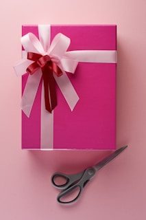 A pink gift box adorned with a large pink and red ribbon bow sits on a matching pink background. Next to the box, a pair of gray scissors is placed with handles facing away.