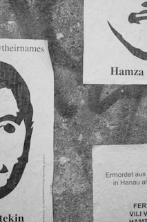 Two printed posters are placed against a rough, textured wall. The left poster features a stylized portrait with the partial text 'theirnames' and possibly a name below. The right poster includes a partial face image with the word 'Hamza' and text that appears to be in German. The images are in black and white.