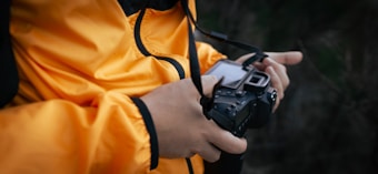 A person wearing a bright orange jacket holds a black digital camera, possibly adjusting settings or preparing to take a photograph. The camera strap is visible, and the background is blurred, suggesting an outdoor setting.