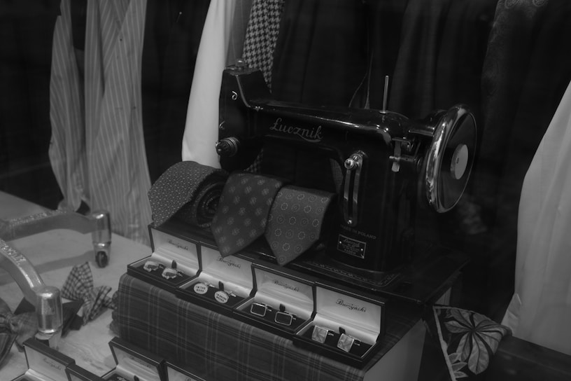 A vintage sewing machine displayed among neatly folded ties and boxes, with various shirts hanging in the background. The image is in black and white, showcasing a classic and elegant setting typically found in a tailor's or clothing store window.