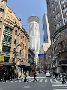 A bustling cityscape with a mix of architectural styles, featuring a prominent modern skyscraper with a unique UFO-like structure at its top, surrounded by older brick and concrete buildings. People are walking through a busy intersection under clear skies. Street signs, power lines, and various storefronts, including a restaurant and spa, are visible.