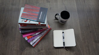 A stack of well-used notebooks and pens next to a steaming cup of tea by a window.