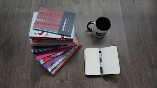A stack of diverse books with a pen and notebook on a wooden table.