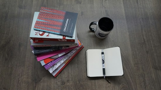 A stack of books is arranged on a wooden floor, alongside an open notebook with a pen resting on it. Nearby, a black and white mug is also placed on the floor.