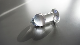 A transparent glass object with a rounded top and flat base rests on a reflective surface. The object casts a soft shadow and reflects light, creating subtle highlights and gradients. The background is a plain, smooth grey surface with streaks of light crossing diagonally.