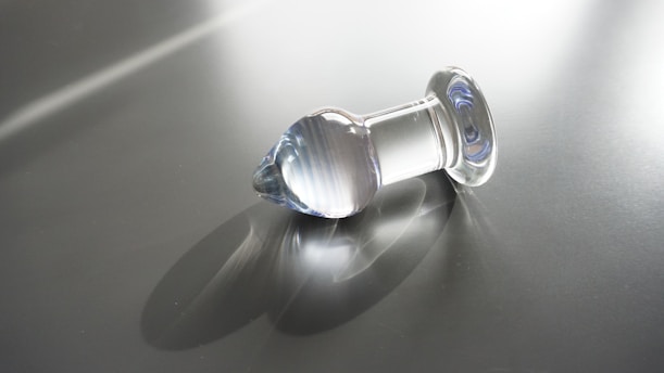 A transparent glass object with a rounded top and flat base rests on a reflective surface. The object casts a soft shadow and reflects light, creating subtle highlights and gradients. The background is a plain, smooth grey surface with streaks of light crossing diagonally.