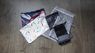 A collection of various fabric pouches are laid on a wooden floor. The pouches have different designs and colors, including a colorful patterned one, a gray mesh-like one, and several others with simple, muted patterns.