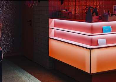 A counter with a modern, geometric design features gradient lighting in shades of pink and orange. The background includes small tiled walls, and there are several items on the counter, including a blue rectangular object and a small container with bottles.