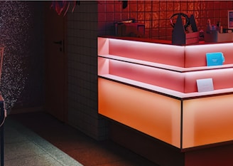 A counter with a modern, geometric design features gradient lighting in shades of pink and orange. The background includes small tiled walls, and there are several items on the counter, including a blue rectangular object and a small container with bottles.