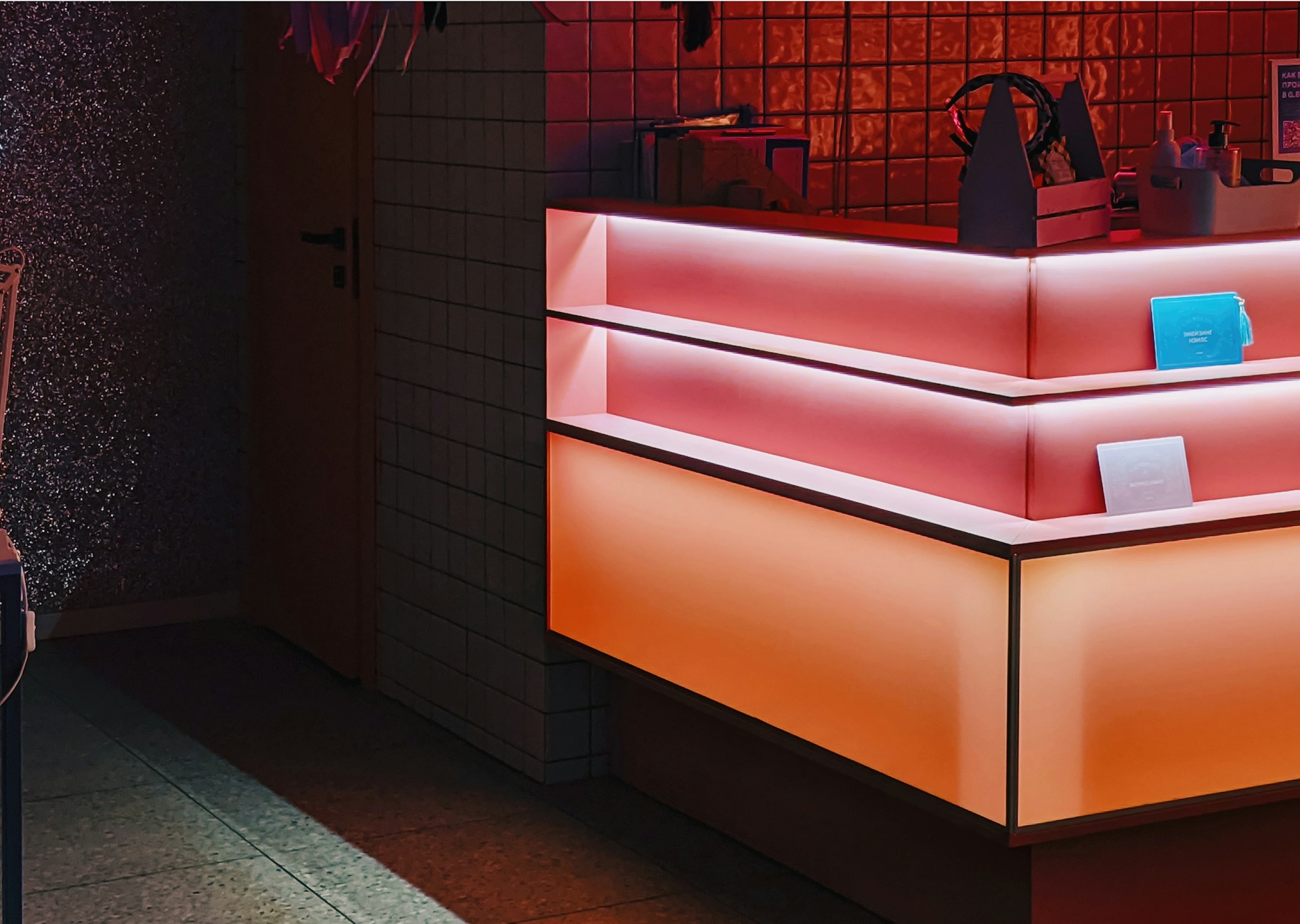 A counter with a modern, geometric design features gradient lighting in shades of pink and orange. The background includes small tiled walls, and there are several items on the counter, including a blue rectangular object and a small container with bottles.