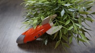 A small, red toy rocket with white fins is nestled among lush green leaves on a wooden surface. The pointed nose of the rocket is gray, and there are small black circular windows on its side. The setting gives a playful juxtaposition of nature and technology.