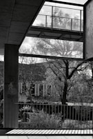 A black and white photograph featuring an urban outdoor scene with a modern architectural structure in the foreground. A tree with bare branches is visible in the background, casting intricate shadows on the ground. A railing with vertical bars is prominently positioned, with sunlight casting linear patterns. Graffiti, including a mushroom and the words 'Happy Life,' is painted on a pillar.